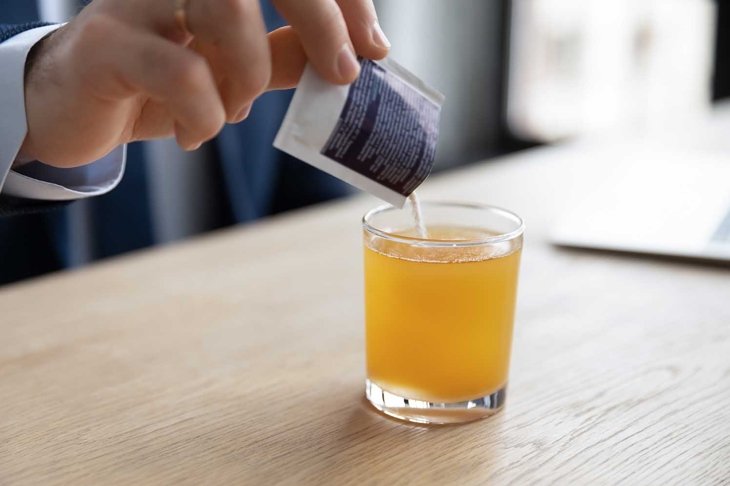 Close up young businessman pouring soluble anti-influenza orange powder with citrus taste in glass of water, feeling first grippe illness symptoms at workplace or relieving headache with painkiller.